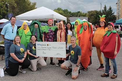 Group of people, some in fruits and veggies costumes, stand with large check at Douglas County farmers market