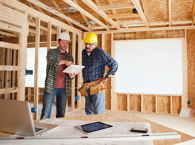 Two construction workers in hard hats discuss plans in room with unfinished walls