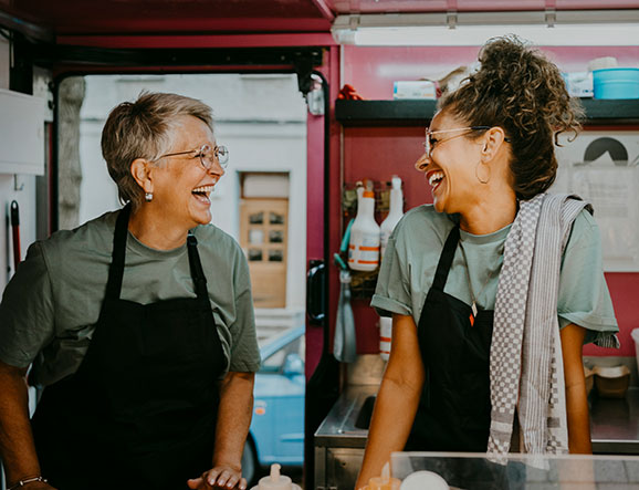 Two restaurant employees wearing black aprons laugh together in kitchen