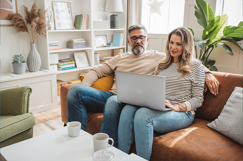 Middle aged couple sit together on orange couch in living room looking at laptop