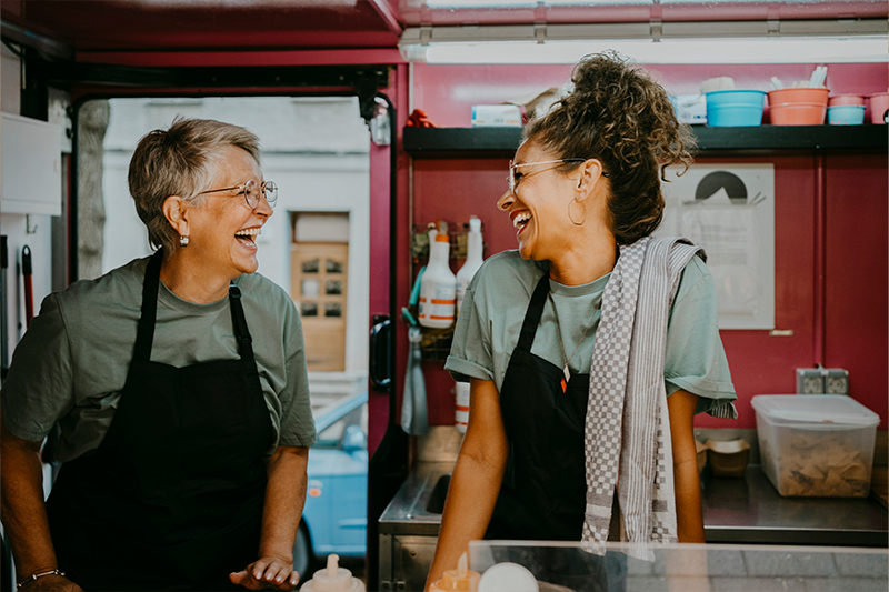 two restaurant workers wearing aprons laugh together in kitchen