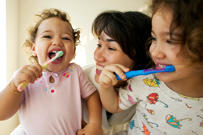 Smiling parent helps two young children brush teeth