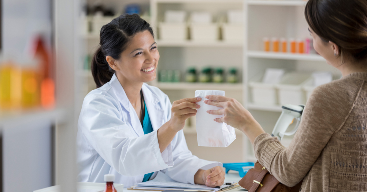 A smiling pharmacist hands a prescription to a patient.