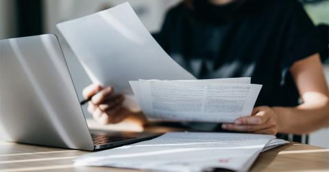 A person holds papers and a pen while sitting at a notebook computer.