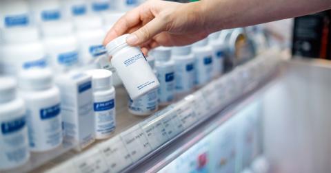 A pharmacist holds a prescription bottle above a shelf full of prescription bottles